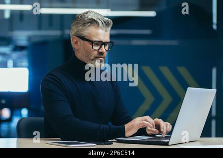 Serious mature gray-haired businessman working inside modern office, man thoughtfully typing on laptop at workplace. Stock Photo