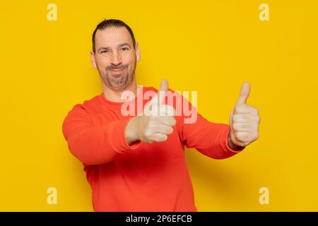 Portrait of a cheerful bearded hispanic man in red sweatshirt smiling and showing thumbs up to camera isolated over yellow background. Stock Photo