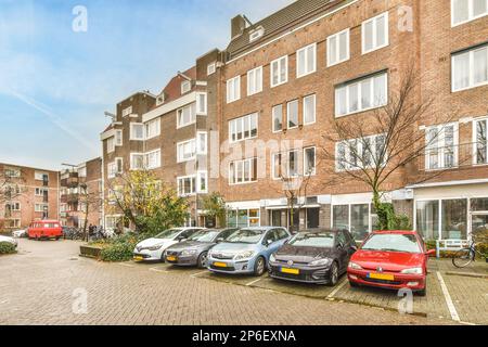 several cars parked on the side of a street in front of a brick building with many windows and bales Stock Photo