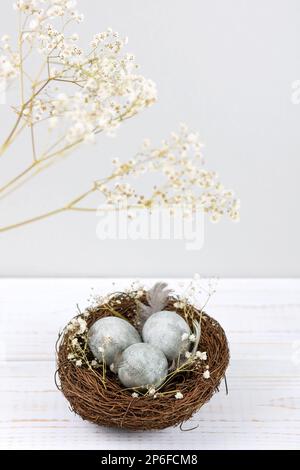 A brown nest of twigs with three gray Easter eggs, feathers and sprigs of gypsophila on a white wooden table. Minimalistic Easter concept Stock Photo
