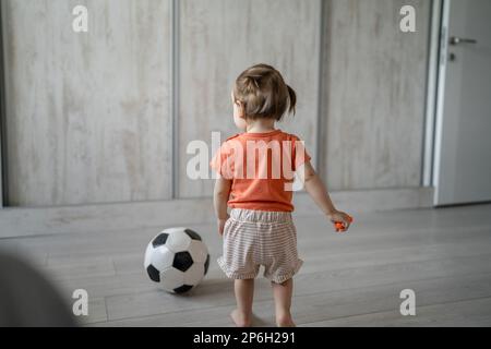 back view of one girl small caucasian toddler daughter wearing orange shirt while walking or standing by the football ball growing up development spor Stock Photo
