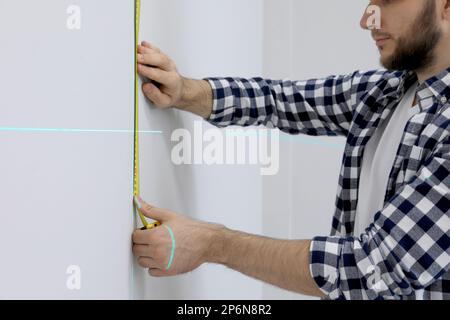 Man using cross line laser level and tape for accurate measurement on light wall, closeup Stock Photo
