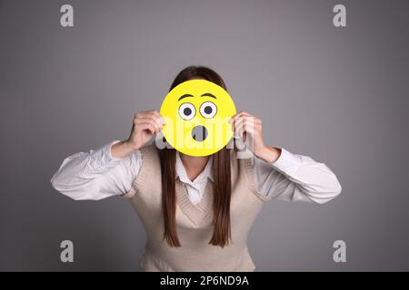 Woman covering face with shocked emoji on grey background Stock Photo