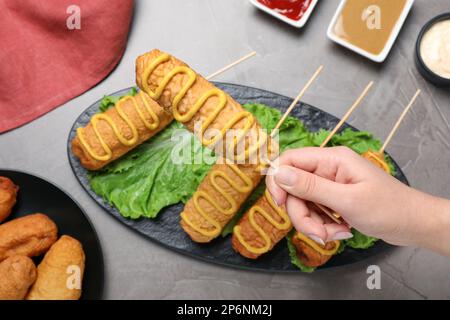 Woman holding delicious corn dog with mustard at grey table, top view Stock Photo