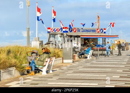 people sitting at an ice cream stand on the beach with flags flying in the sky and sand dunes behind them Stock Photo