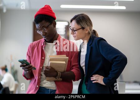 Two diverse students standing in classroom using smartphone during study break Stock Photo