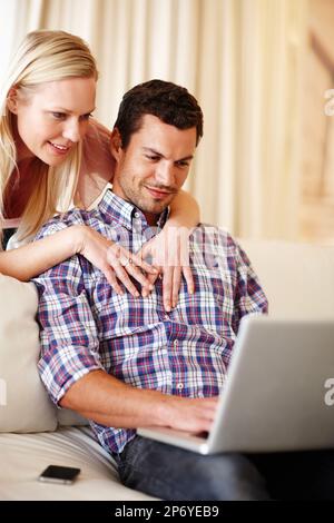 Keeping in touch with friends. A beautiful woman hugging her husband from behind while he sits on the couch working on his laptop. Stock Photo