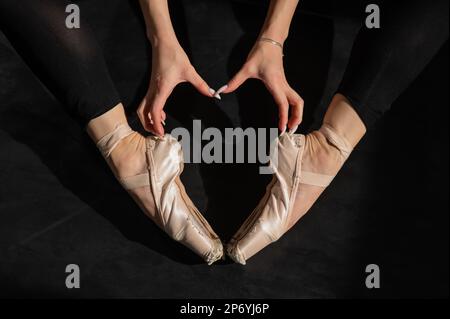 A faceless ballerina sits on the floor and makes a heart shape with her hands and pointe shoes. Top view.  Stock Photo