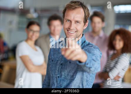You could be our future colleague. Smiling man pointing at you a friendly group of office staff in the background. Stock Photo