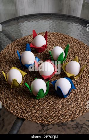 photo white eggs lying in a multicolored container on a wicker napkin on a glass table Stock Photo