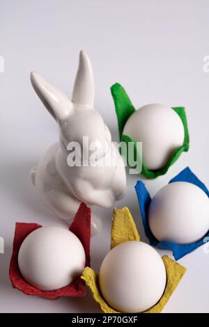 photo white chicken eggs in colored individual packages and a white ceramic rabbit on a white background Stock Photo