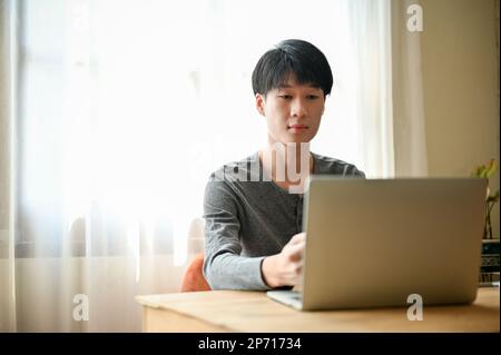 Concentrated young Asian man in causal clothes using laptop computer at a desk in his home living room. Stock Photo