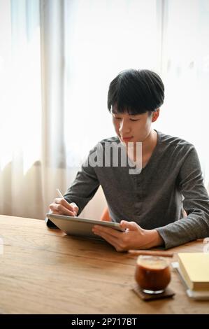 Handsome young Asian man in casual clothes using digital tablet, writing or noting something on his tablet while relaxing in the cafe. Stock Photo