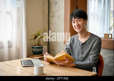 Happy and smiling young Asian man in casual clothes sits in the beautiful cozy coffee shop with his book and portable laptop. Stock Photo