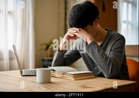 Stressed and depressed young Asian man is reading a book in the coffee shop co-working space, preparing for the exam. Stock Photo