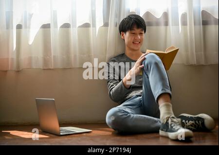 Happy and cute young Asian man in casual clothes sits on the floor in his room and enjoys reading a book. leisure or hobby concept Stock Photo