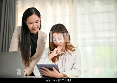 Positive and successful millennial Asian businesswoman looking at tablet screen, thinking and planning a project with her female assistant in the offi Stock Photo