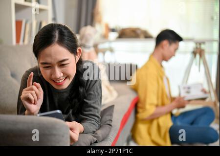 Happy and joyful young Asian woman is laughing, watching something funny on her phone, and laying on the sofa while her boyfriend sits in the back. Stock Photo