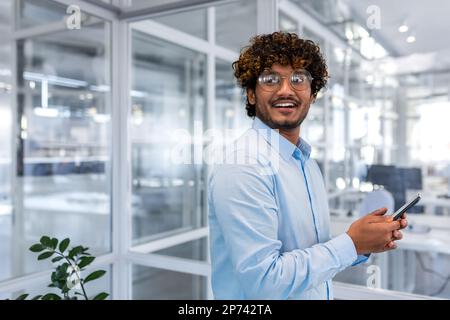 Portrait of successful hispanic programmer engineer developer inside office, man in shirt and glasses holding smartphone in hands smiling and looking out window, businessman at workplace young happy. Stock Photo