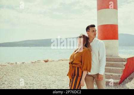 A couple stands near a lighthouse on the beach against the backdrop of the sea and mountains. Vacation love, honeymoon, travel. Stylish couple laughin Stock Photo
