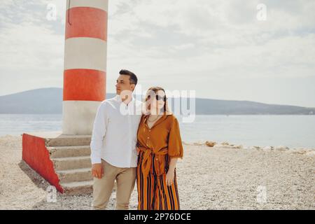A couple stands near a lighthouse on the beach against the backdrop of the sea and mountains. Vacation love, honeymoon, travel. Stylish couple laughin Stock Photo