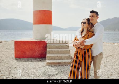 A couple stands near a lighthouse on the beach against the backdrop of the sea and mountains. Vacation love, honeymoon, travel. Stylish couple laughin Stock Photo