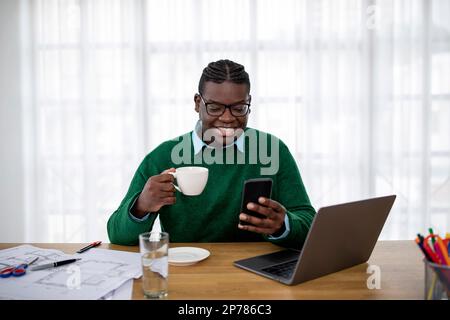 Happy Black Businessman Using Phone Texting Drinking Coffee At Workplace Stock Photo