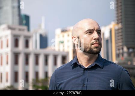 Serious attractive man standing against a cityscape and thinking. Bald Caucasian guy feeling depressed, alone on a city street Stock Photo