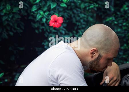 Thoughtful sad man looking down, sitting in the forest. Bald Caucasian man on a green foliage background. Anxiety, depression state of mind concept Stock Photo