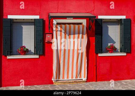 doorway with curtain and windows with shutters on bright red coloured house at Burano, Venice, Italy in February Stock Photo