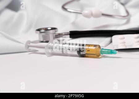 Close-up of a syringe loaded with medication. in the background: pills, stethoscope and doctors gown Stock Photo