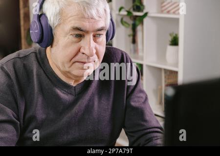 Cheerful older person with headphones sitting at table at cozy room and working on computer. Old Man using laptop, watching online program, funny vide Stock Photo