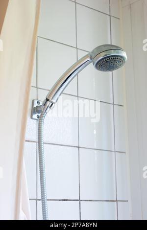 Shower head and cord hanging on the bathroom tile wall next to shower curtain. Stock Photo