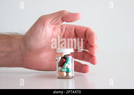 Caucasian male hand reaching out for a glass bottle full of various colored medical pills, tablets and capsules. Close up studio shot, isolated on whi Stock Photo