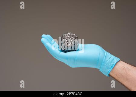 New kitchen metal wire scrubber held in hand by Caucasian male hand wearing a blue latex glove. Close up studio shot, isolated on gray background. Stock Photo
