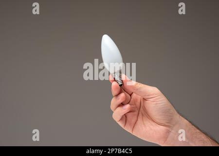 White mate incandescent light bulb held in hand by Caucasian male hand. Close up studio shot, isolated on gray background. Stock Photo