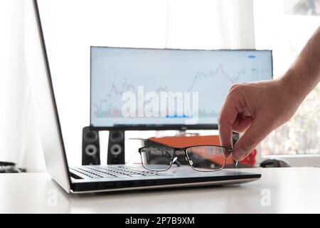 Clear reading glasses, black rim, on a laptop keypad. Blurred large computer monitor in the background showing stock market graph. Male hand grabbing Stock Photo