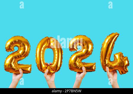 hands holding some golden number-shaped balloons forming the number 2024, as the new year, on a blue background with some blank space on top Stock Photo