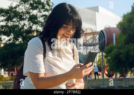 happy young venezuelan immigrant medical student in argentina, sitting in the university park, using the phone surfing the internet on break. Stock Photo