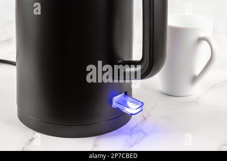 Electric kettle switch glows blue while turned on. Matte black kettle for heating water to boiling for tea and coffee. Modern class 1 electrical appli Stock Photo