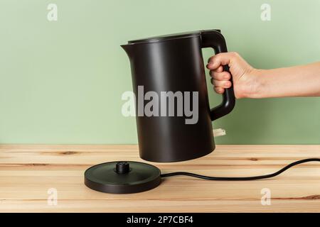 Matte black kettle kettle in a woman hand. Person hand taking off electric kettle from power base on a wooden countertop. Class 1 electrical appliance Stock Photo