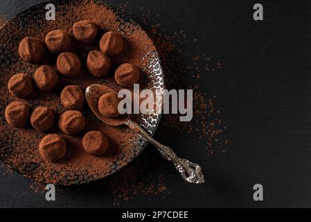 Chocolate truffle balls and spoon on a black plate. Homemade dark chocolate candies sprinkled with cocoa powder. Sweet food concept. Copy space. Stock Photo