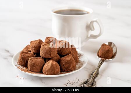 Chocolate truffle balls on a white plate. Heap of dark chocolate candies in cocoa powder, cup of coffee and spoon on a marble table. Sweet food. Stock Photo