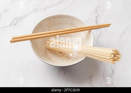 Dried udon nudles in a bowl closeup. Bunch of raw japanese vegan pasta from wheat flour and bamboo chopsticks over bowl against marble countertop. Stock Photo