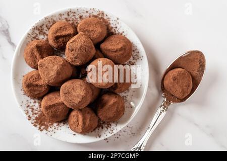 Gourmet chocolate truffles on a white saucer. Plate full of dark chocolate candy balls in cocoa powder and spoon over marble surface. Sweet food. Stock Photo