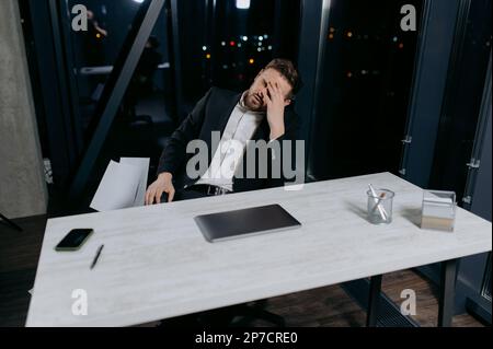 tired and stressed young office worker or businessman works late at desk with his laptop Stock Photo