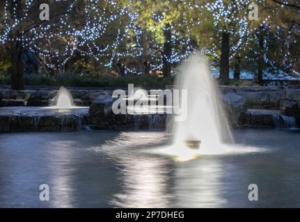 Fountains and pools surrounded by stones underneath trees and fairy lights by night. Long exposure. Stock Photo