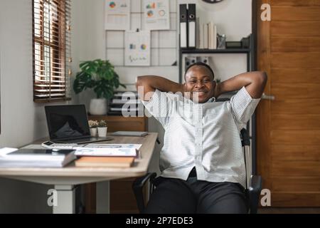 Feeling joy. Cheerful african businessman is reclining on chair and holding his hands behind head while looking up with wide smile Stock Photo