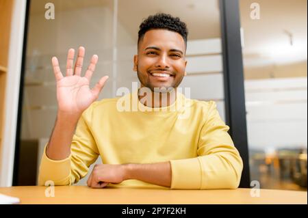 Portrait of smiling and friendly indian male freelancer using laptop for virtual meeting and waving at the camera. Latin guy involved video call, staying in touch with colleagues working remotely Stock Photo