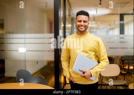 Smiling young indian businessman, arab male student, eastern employee, manager, coworker wearing casual yellow pullover standing carrying laptop, looking at the camera standing in office Stock Photo